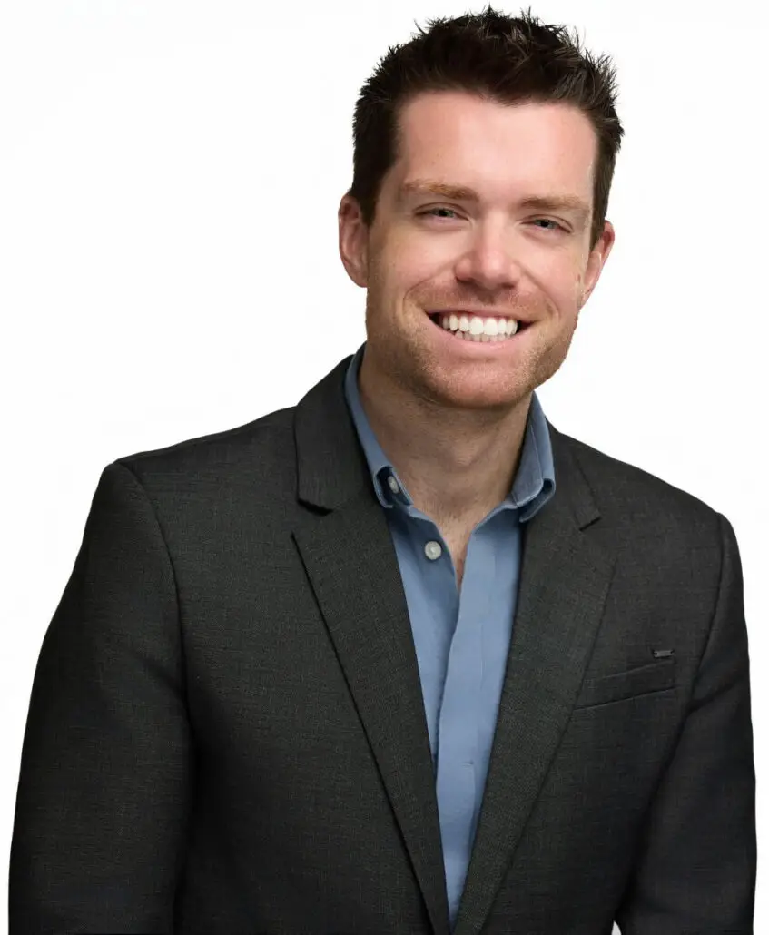 A man with short brown hair wearing a dark blazer over a blue button-up shirt is smiling at the camera, ready to discuss the 2025 Carnival Cruise Director Schedule, against a plain white background.