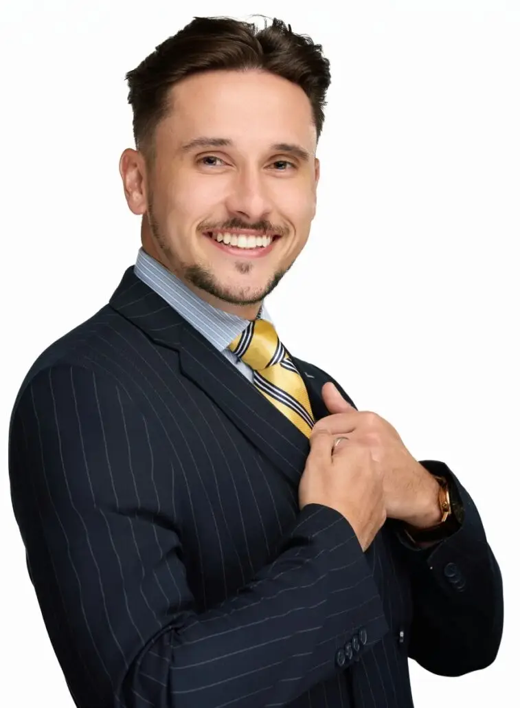 A man with short brown hair and a trimmed beard, dressed in a dark pinstripe suit and yellow striped tie, smiles as he adjusts his jacket. He looks ready to take on the 2025 Carnival Cruise Director Schedule, standing against a plain white background.