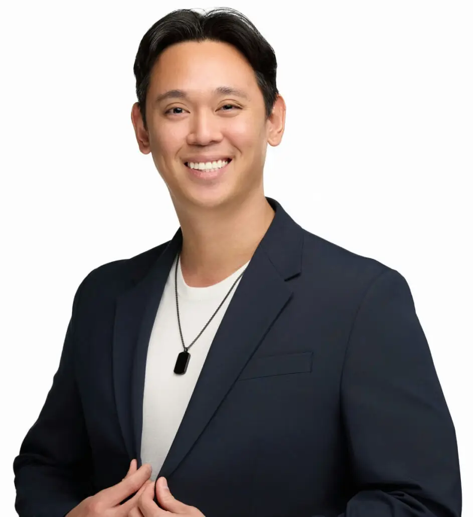 A man wearing a dark blazer over a light-colored shirt and a rectangular pendant necklace stands against a plain white background, smiling at the camera with his hands adjusting his jacket—exuding the confidence of a seasoned cruise director.