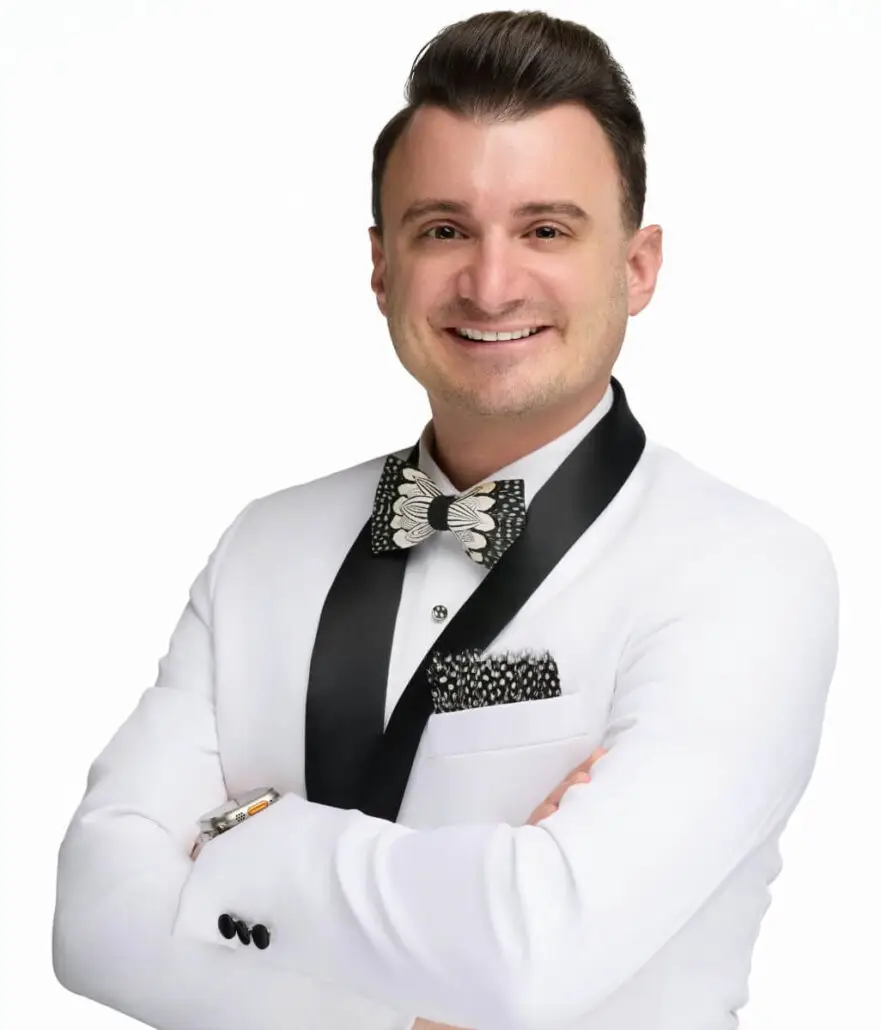 A man with short, dark hair smiles confidently with arms crossed. Dressed in a white tuxedo with black lapels and a patterned bow tie, he exudes the polished style of a cruise director—perfect for the Carnival Cruise Director Schedule.