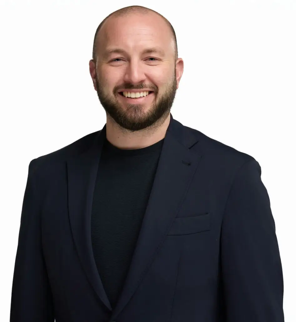 A man with a shaved head and a beard is smiling. He is wearing a dark blazer over a dark shirt, standing against a plain white background, ready to discuss the 2025 Carnival Cruise Director Schedule for your next cruise ship adventure.