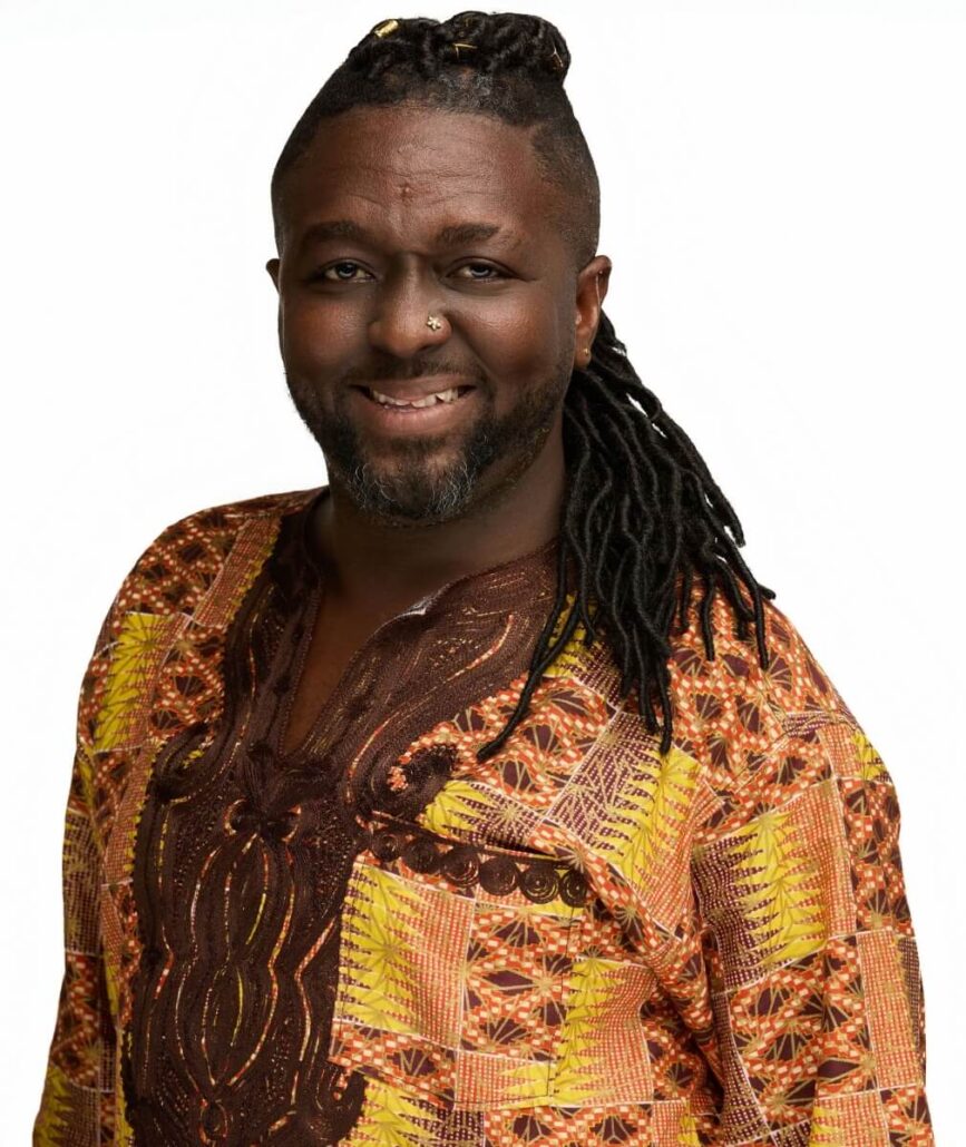 A man with long black dreadlocks and a nose ring smiles. He is wearing a colorful patterned shirt with shades of brown, yellow, and orange—ready for fun, just like the 2025 Carnival Cruise Director Schedule. The background is plain white.