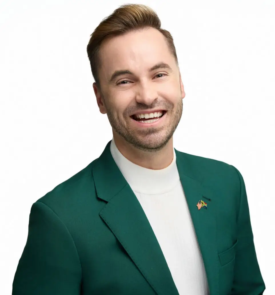 A man with short light brown hair and a beard is wearing a green suit jacket and white turtleneck. He is smiling at the camera against a plain white background, dressed smartly as if ready for the 2025 Carnival Cruise Director Schedule on a cruise ship.