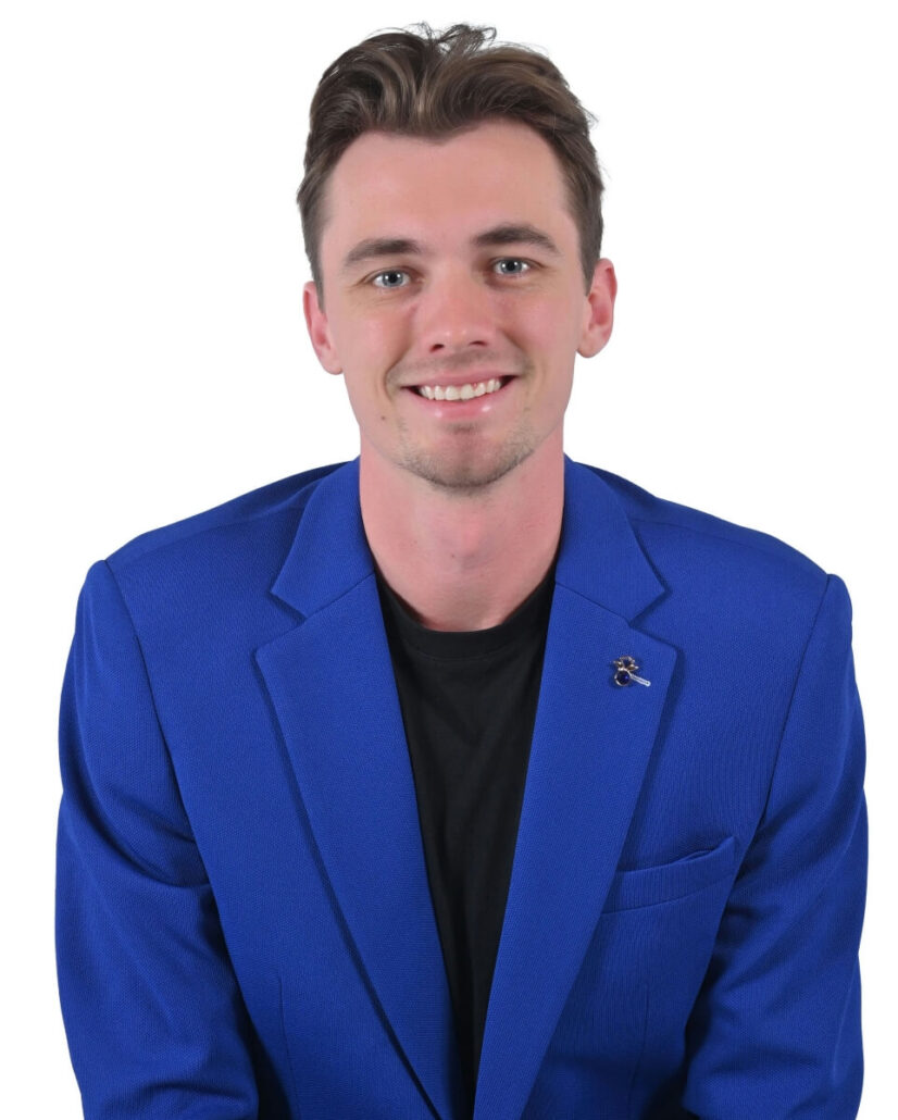A young man with short brown hair is smiling at the camera. Wearing a bright blue blazer over a black shirt, he stands against a plain white background, ready for the 2025 Carnival Cruise Director Schedule aboard your favorite Carnival cruise ship.