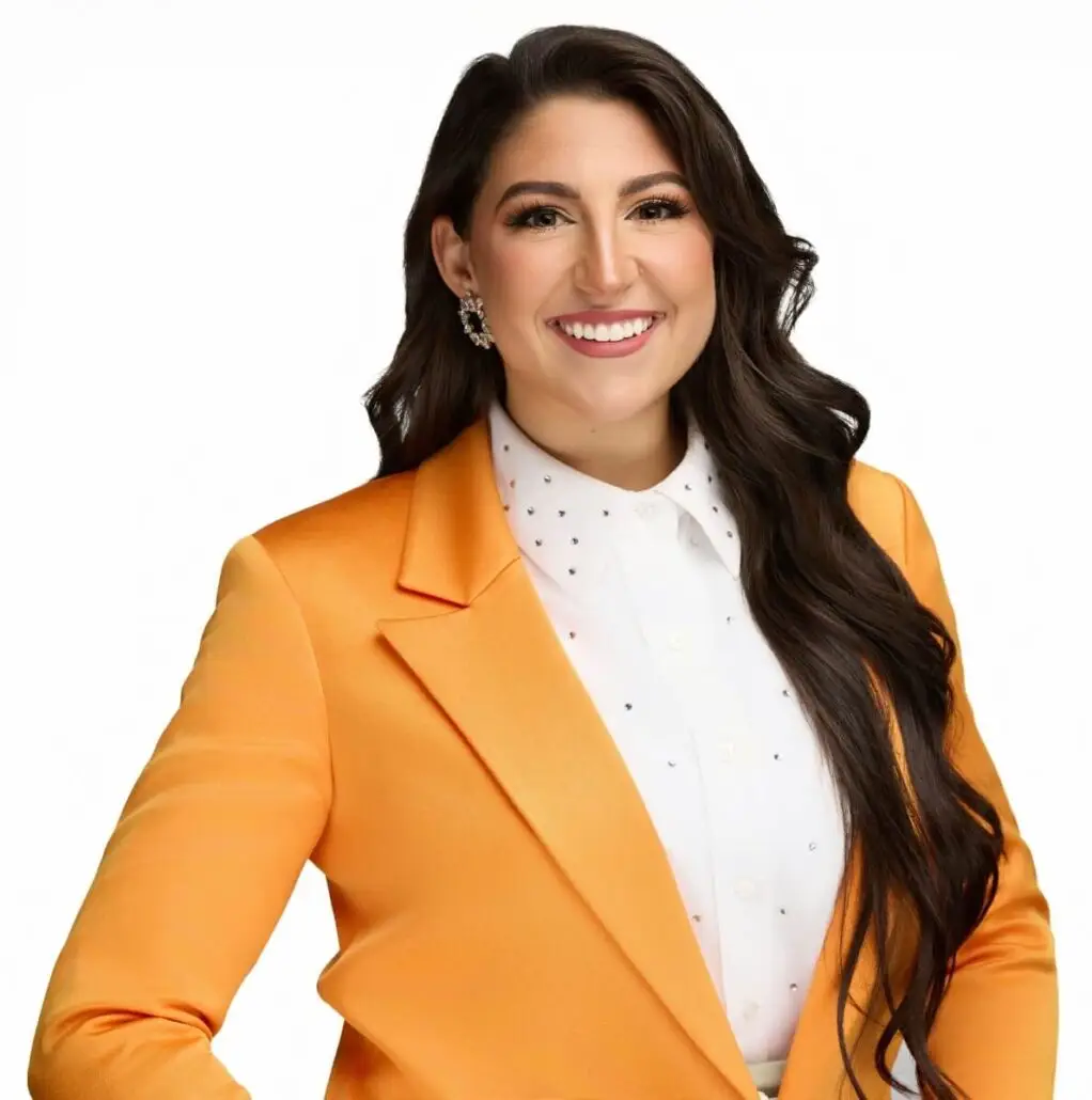 A woman with long dark hair, wearing a bright orange blazer over a white collared shirt with small embellishments, stands smiling against a plain white background, exuding the confident energy of a cruise director ready for the 2025 season.