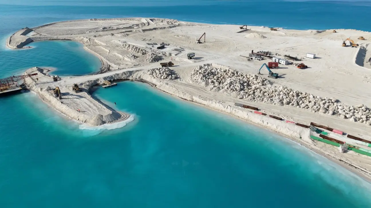 An aerial view of a construction site on a sandy island surrounded by turquoise water, where MSC Cruises is developing land and amenities for an exclusive private island experience for future guests. Heavy machinery and vehicles are scattered around the site.
