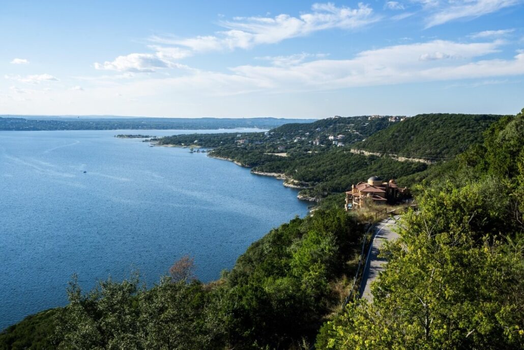 A large Lake Travis bordered by green hills, with scattered houses and Lake Travis boat slips along the shoreline. A winding road is visible in the foreground under a blue sky with scattered clouds.