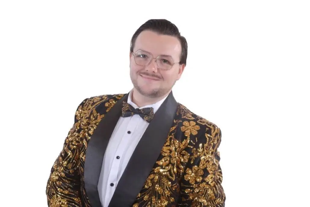 A person wearing glasses, a white dress shirt, and a black and gold floral-patterned suit jacket with a matching bow tie stands against a plain white background—ready to headline the 2025 Carnival Cruise Director Schedule as your cruise director.