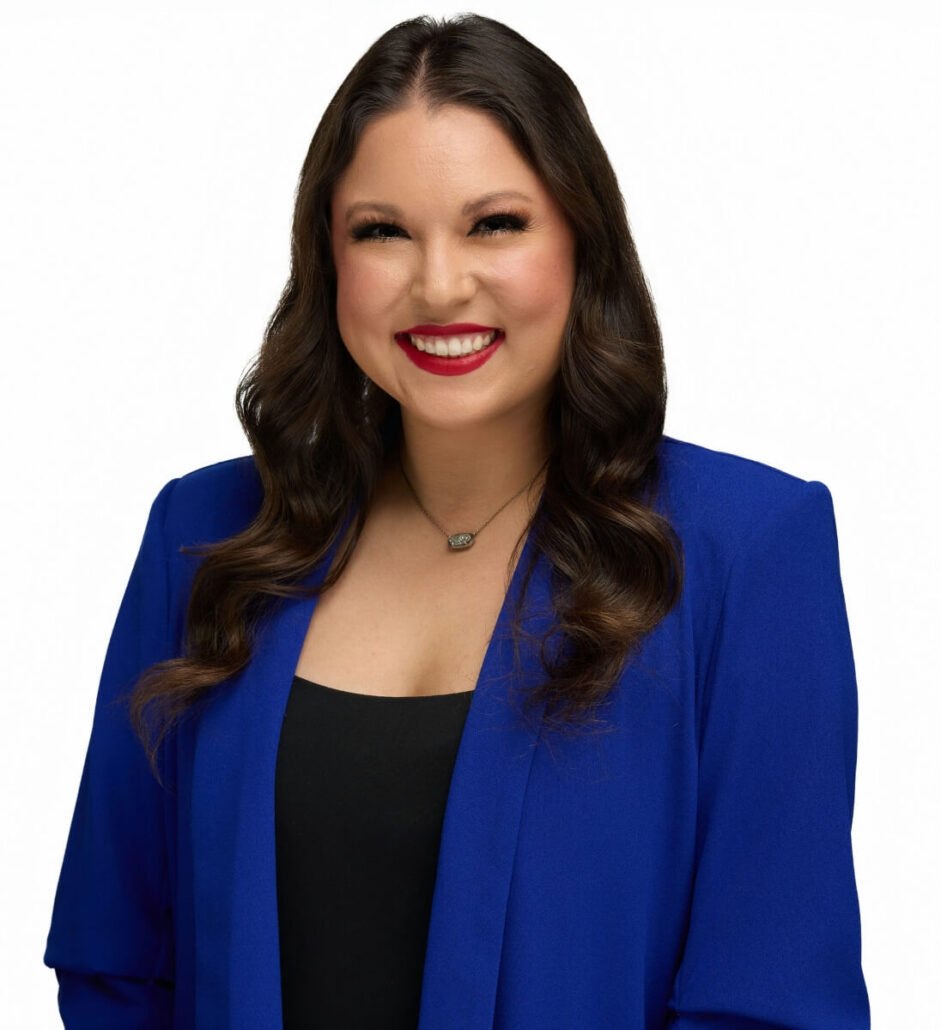 A woman with long, wavy brown hair is smiling. She is wearing a bright blue blazer over a black top and a necklace, looking ready for her role as Cruise Director. The background is plain white.