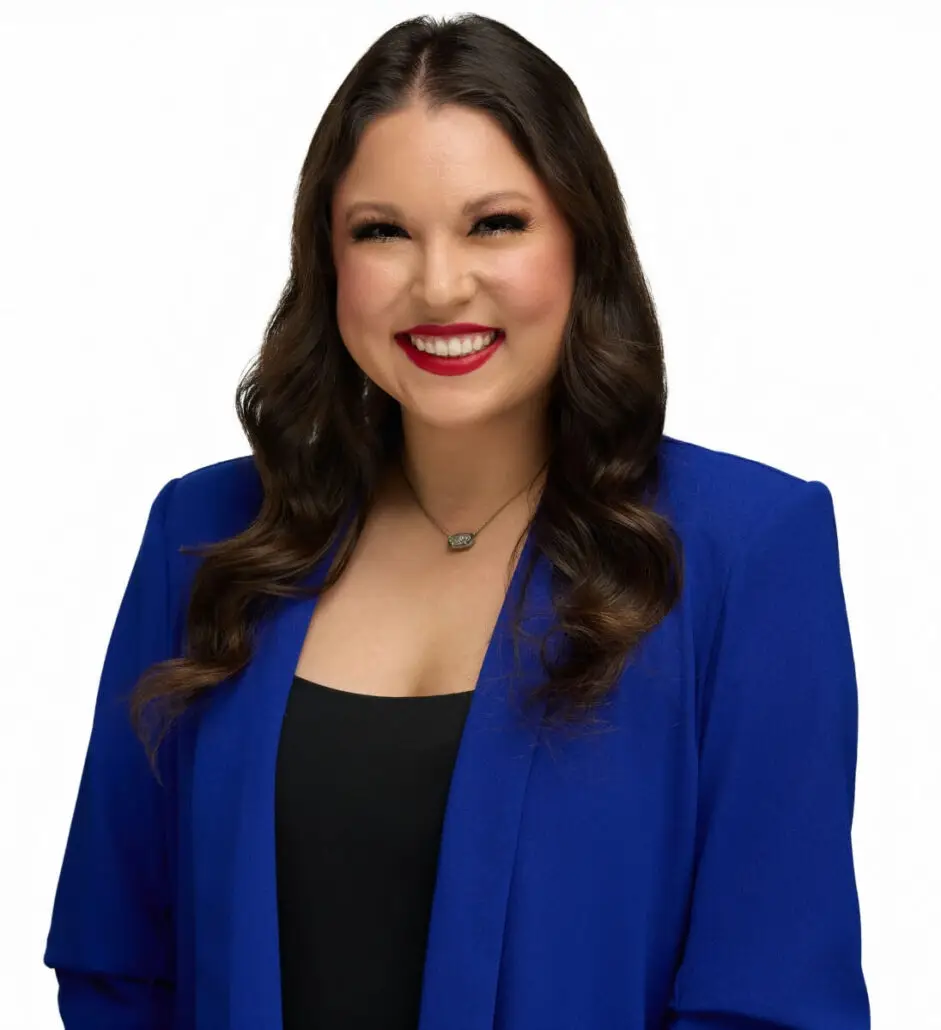 A woman with long, wavy brown hair is smiling. She is wearing a bright blue blazer over a black top and a necklace, looking ready for her role as Cruise Director. The background is plain white.