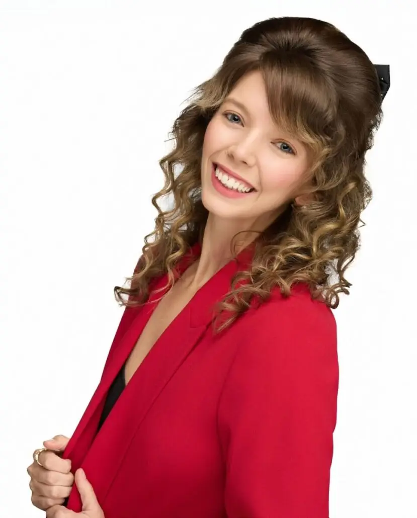 A woman with curly brown hair, wearing a red blazer over a black top, smiles at the camera against a plain white background, ready to share the latest Carnival Cruise Director Schedule for Carnival 2025.