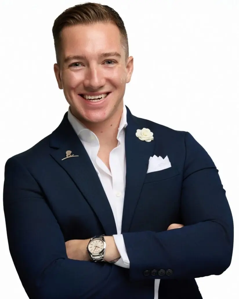 A man with short light brown hair is smiling with arms crossed. Dressed smartly in a navy blue blazer and white shirt, he looks ready for his role as Carnival Cruise Director, perhaps preparing for the 2025 cruise ship schedule, against a plain white background.