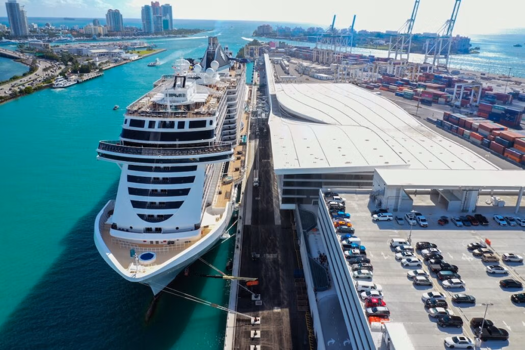 Aerial view of the world's largest cruise ship docked at a Miami port. The ship is beside a bustling cruise terminal, with parked cars lining the area. The sea sparkles turquoise, while cranes and containers dot the distance. Skyscrapers and a bridge enhance the vibrant skyline.