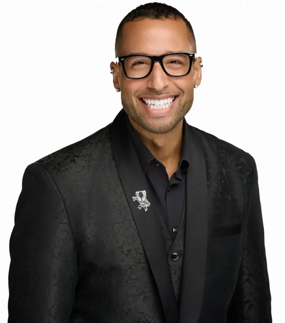 A man wearing glasses and a dark patterned suit jacket over a black shirt smiles at the camera against a white background. He has a pin on his lapel and multiple earrings in his left ear, embodying the style of a professional cruise director.