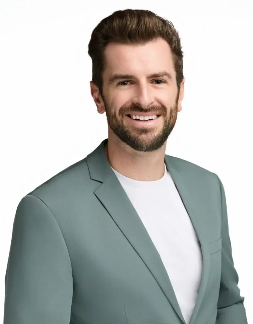 A man with brown hair and a beard is smiling at the camera. Dressed in a light teal blazer over a white t-shirt, he could easily pass for a cruise director ready to announce the 2025 Carnival Cruise Director Schedule. The background is plain white.
