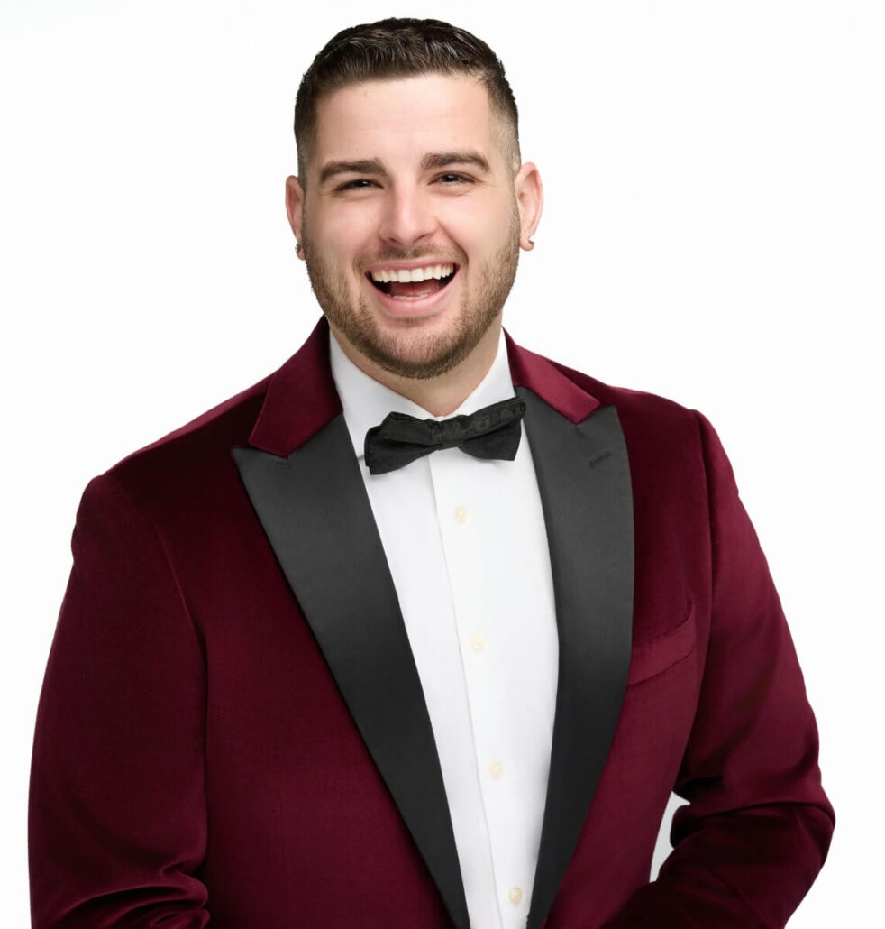 A man with short hair and a beard, dressed in a burgundy suit jacket, white dress shirt, and black bow tie, smiles at the camera against a plain white background—just like one of the cruise directors you might spot on the Carnival Cruise Director Schedule for 2025.