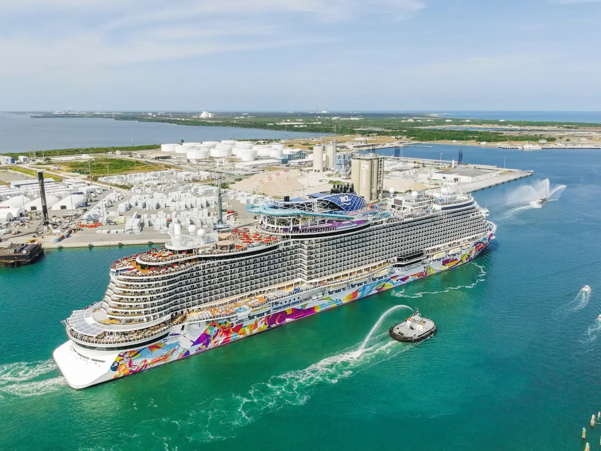 The Norwegian Aqua, a large multi-deck cruise ship adorned with colorful hull artwork, is docked in Port Canaveral’s harbor, guided by a tugboat. Industrial tanks and buildings line the shore under a partly cloudy sky.