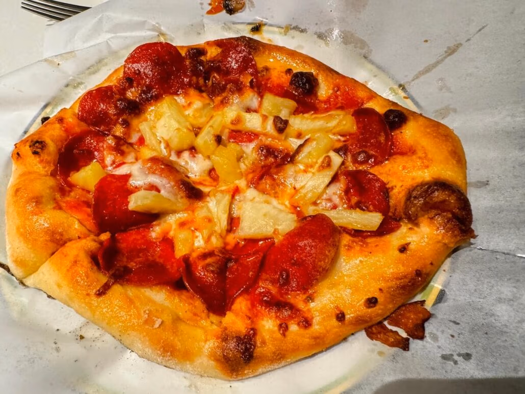 A baked pizza with pepperoni slices and pineapple chunks on melted cheese and tomato sauce, served on white parchment paper aboard the Koningsdam during a scenic Alaska cruise.