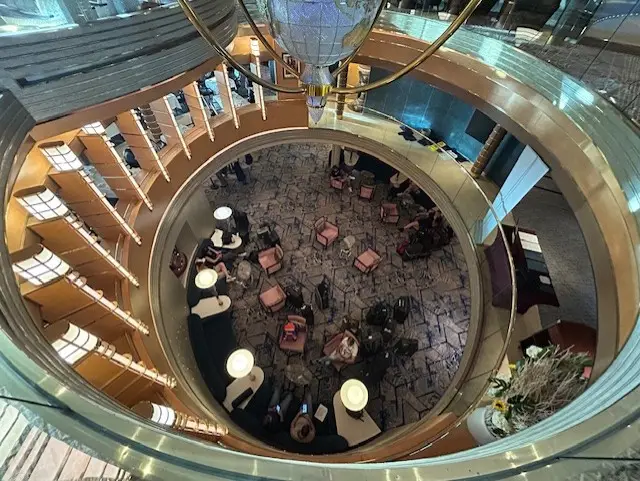 A view from above of Oosterdam’s circular, multi-story atrium with gold railings, seating areas with chairs and tables on a patterned carpet, and people sitting and conversing throughout the space.