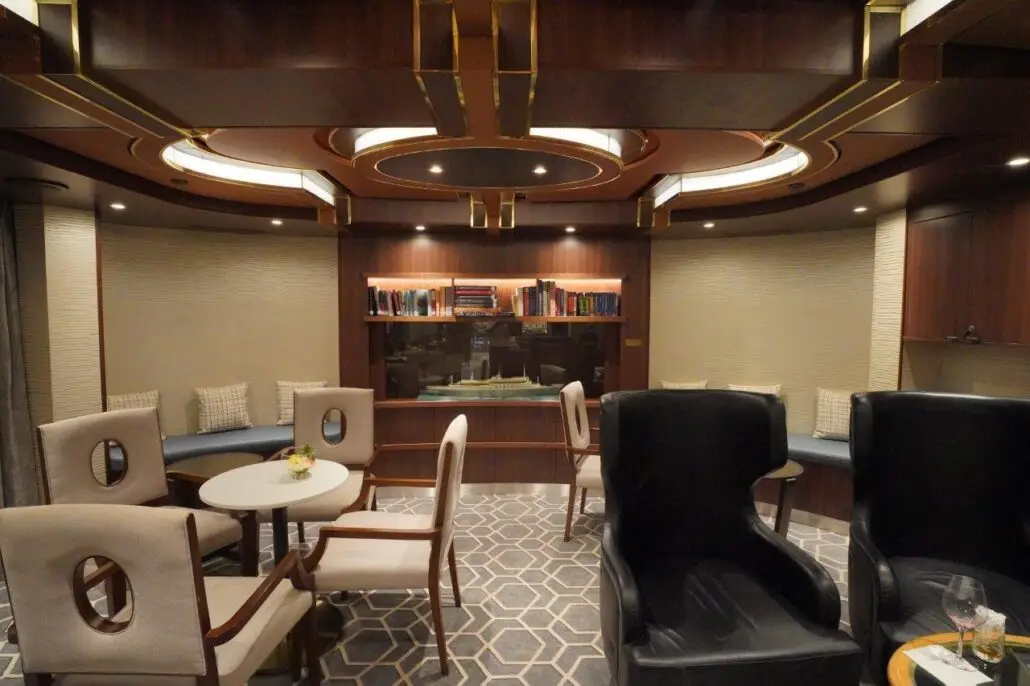 A lounge on the Oosterdam with round tables, cushioned chairs, two high-backed armchairs, built-in bookshelves, a windowside bench with pillows, and geometric carpet under warm ceiling lights—perfect for relaxing after the spa or casino.