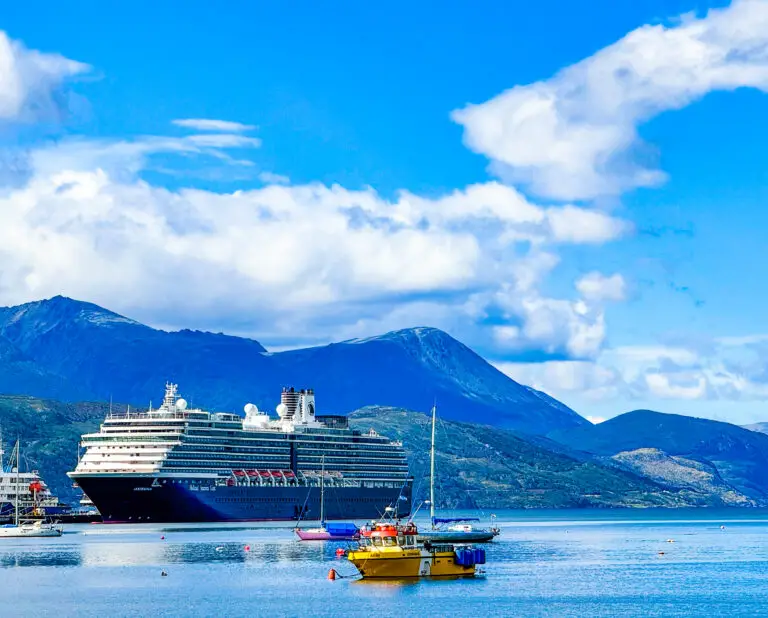 The Oosterdam, a large cruise ship, is docked near the shore with mountains in the background. A small yellow boat and a sailboat float on calm blue water under a partly cloudy sky, perfect for a relaxing spa day onboard.