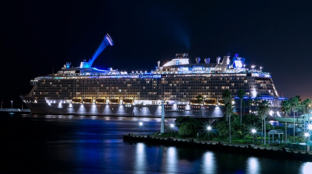 A large, illuminated cruise ship is docked in a harbor at night, with bright blue and white lights reflecting on the water. Trees and lights are visible in the foreground on the shore.