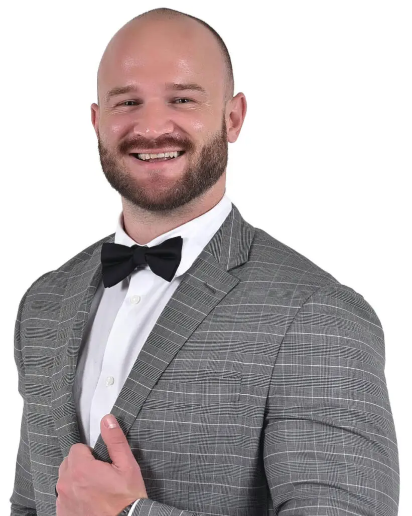 A man with a bald head and beard is smiling while wearing a gray plaid suit, white dress shirt, and black bow tie. Posing confidently like a 2025 Carnival Cruise Director, he holds one side of his jacket against a plain white background.