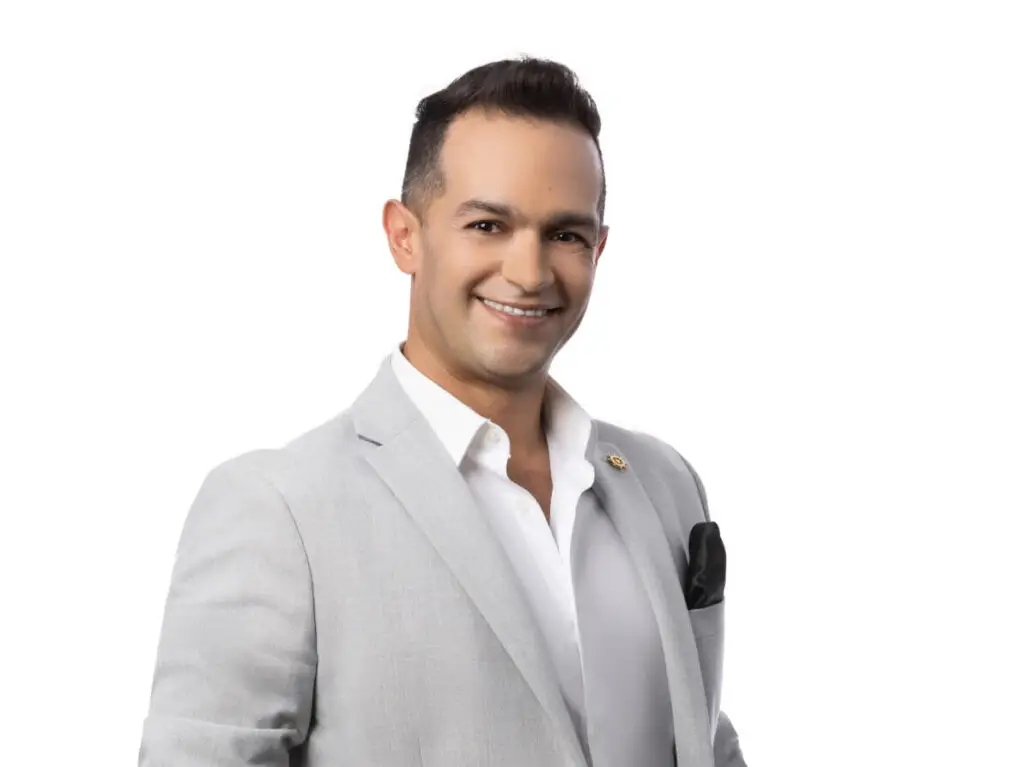 A man with short dark hair wearing a light gray suit, white dress shirt, and black pocket square stands against a plain white background, smiling at the camera—ready to discuss the Carnival Cruise Director Schedule for 2025.