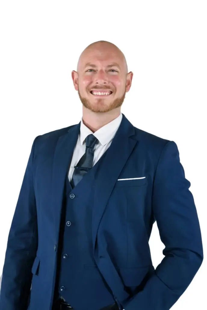 A man in a blue suit, white shirt, and dark tie stands smiling against a plain white background. Bald with a trimmed beard, he poses with one hand in his pocket—ready for his 2025 debut as a Carnival Cruise Director.