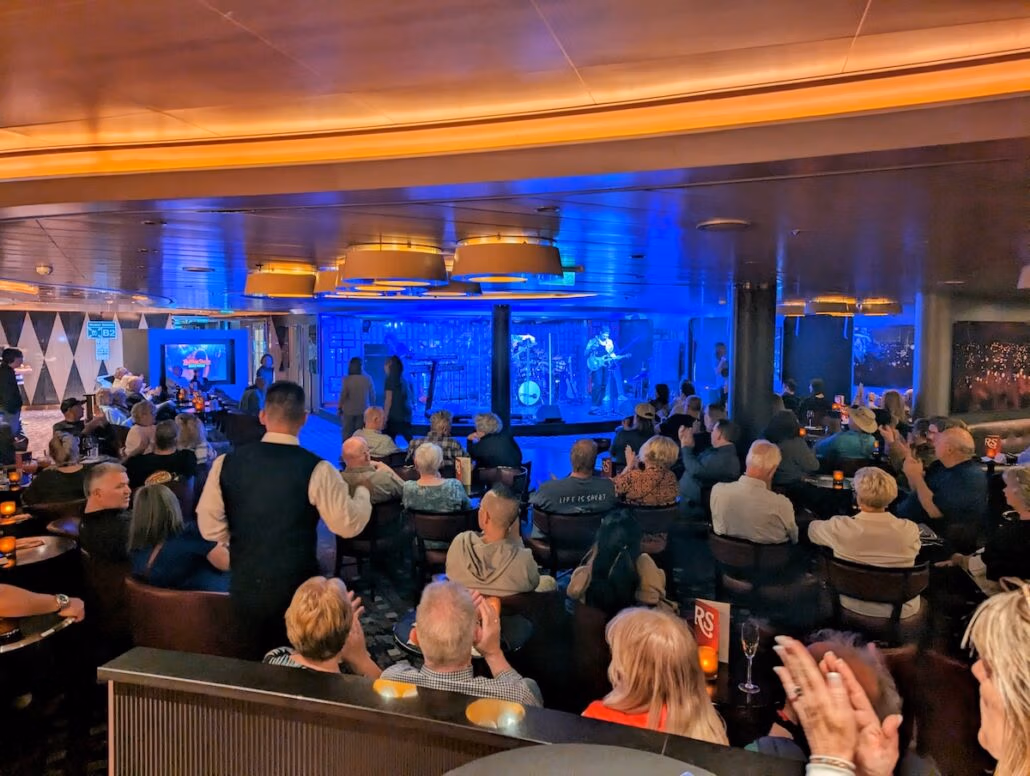 A crowd of people sits in a lounge aboard the Koningsdam, facing a stage where a live band performs under blue lighting. A waiter serves drinks among the audience, and several people are clapping on this elegant Holland America Alaska cruise.