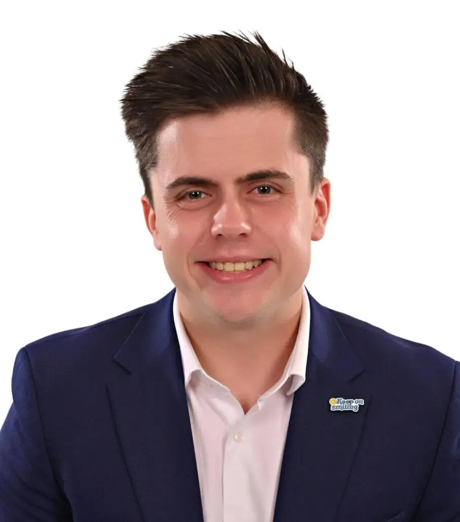 A man with short brown hair, wearing a navy blue suit jacket, white shirt, and a small rectangular pin, smiles at the camera against a plain white background—perfect for a 2025 Carnival Cruise Director Schedule or cruise director profile.