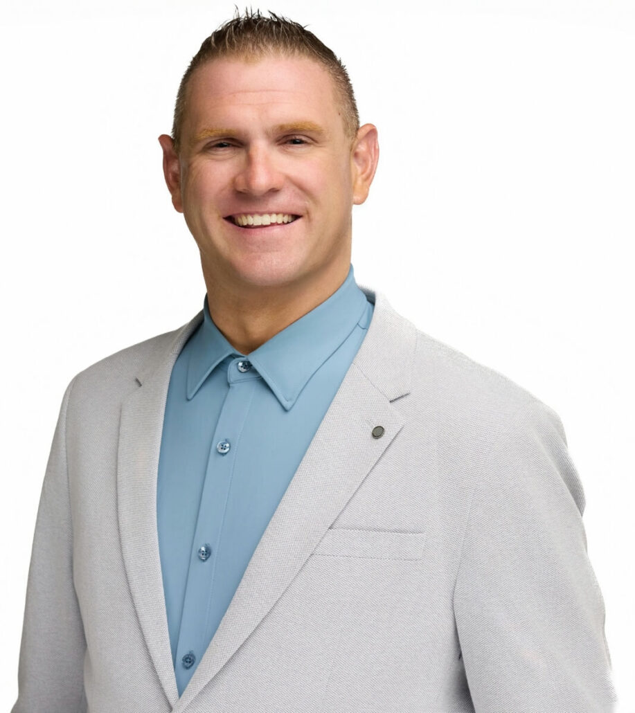 A man with short, light brown hair is wearing a light grey blazer over a buttoned-up blue shirt. He is standing against a plain white background and smiling at the camera, ready to announce the Carnival Cruise Director Schedule for 2025.