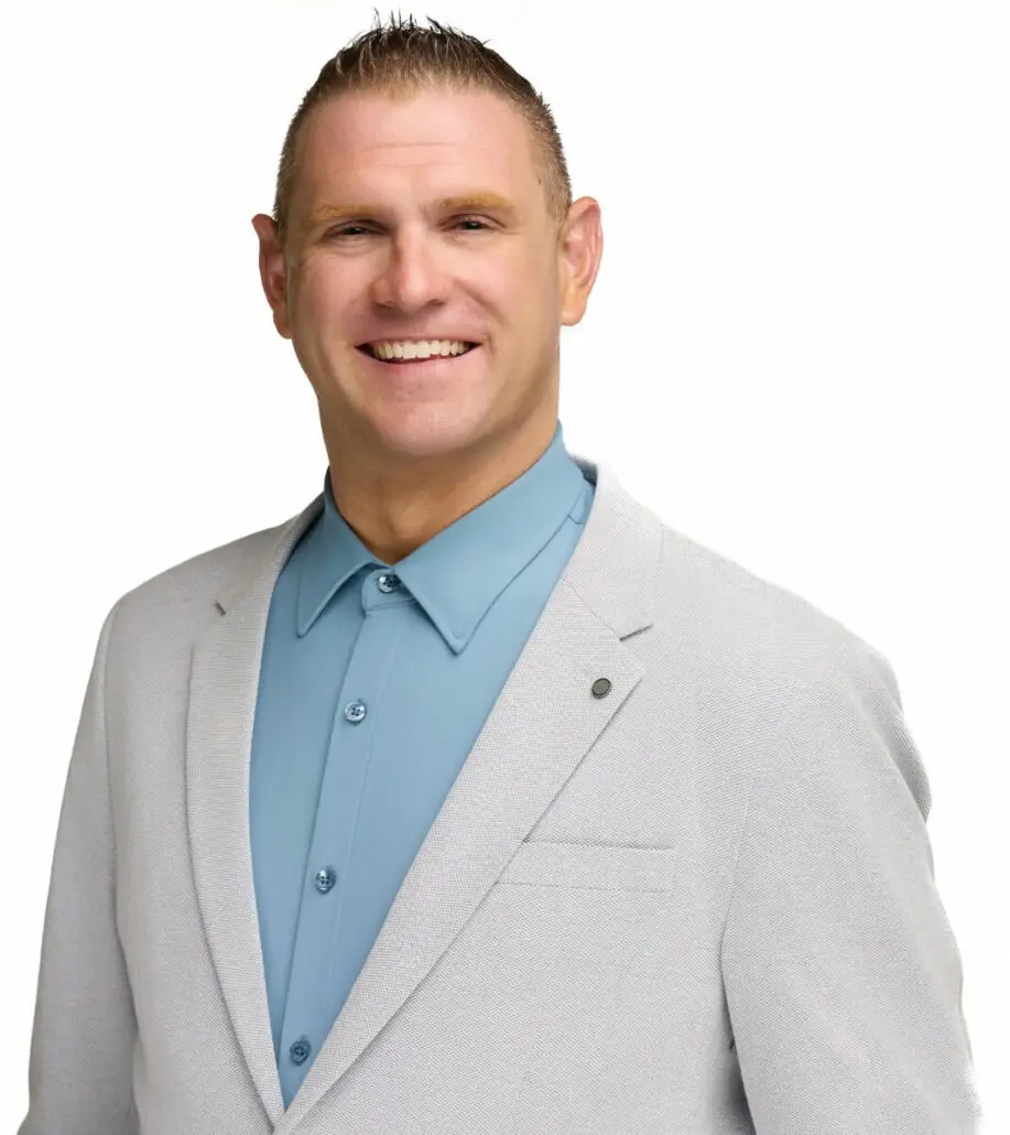 A man with short, light brown hair is wearing a light grey blazer over a buttoned-up blue shirt. He is standing against a plain white background and smiling at the camera, ready to announce the Carnival Cruise Director Schedule for 2025.