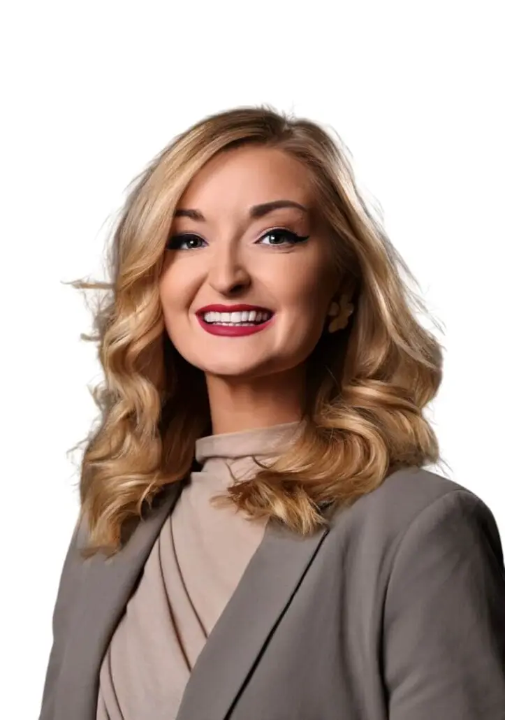 A woman with shoulder-length blonde hair, dressed in a tan blouse and gray blazer, smiles at the camera—ready to discuss the 2025 Carnival Cruise Director Schedule against a plain white background.