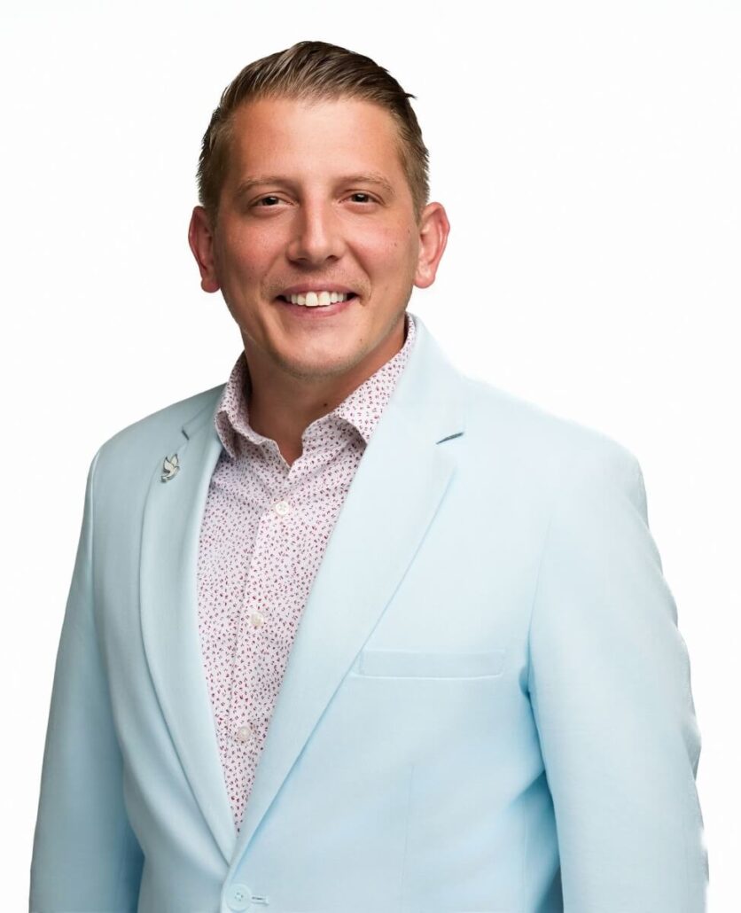 A man with short, slicked-back blond hair smiles at the camera. He is wearing a light blue blazer over a white shirt with a small red pattern, ready to discuss the Carnival Cruise Director Schedule for Carnival 2025. The background is plain white.