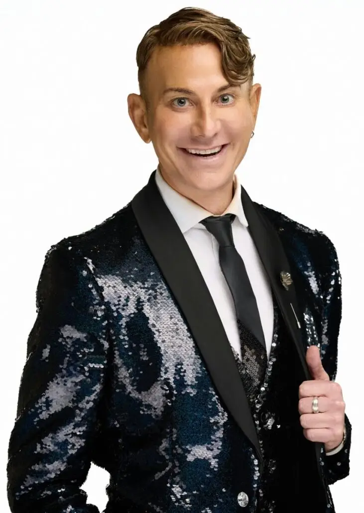 A person with short, styled brown hair is wearing a black sequin jacket, white shirt, black vest, and black tie. Smiling and holding their lapel, they exude the polished look of a Carnival Cruise Director ready for the 2025 schedule. The background is plain white.