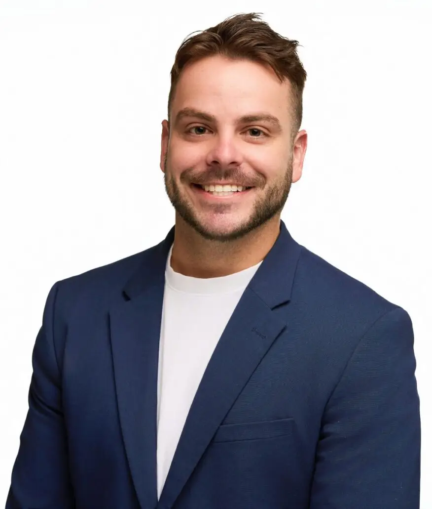 A man with short brown hair and a trimmed beard, dressed in a navy blue blazer over a white t-shirt, smiles at the camera against a plain white background—ready for his 2025 role as Cruise Director.
