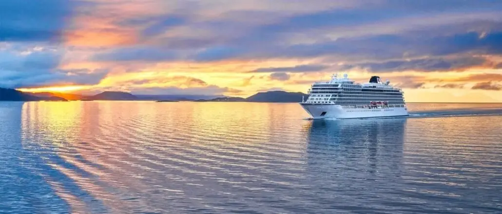 The Viking Polaris, a large white cruise ship, sails on calm water at sunset with passengers aboard, mountains in the distance, and a sky painted in orange, yellow, and blue hues.