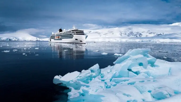 The Viking Polaris, carrying passengers, sails through icy, calm waters surrounded by floating ice and snow-covered mountains under a cloudy sky.