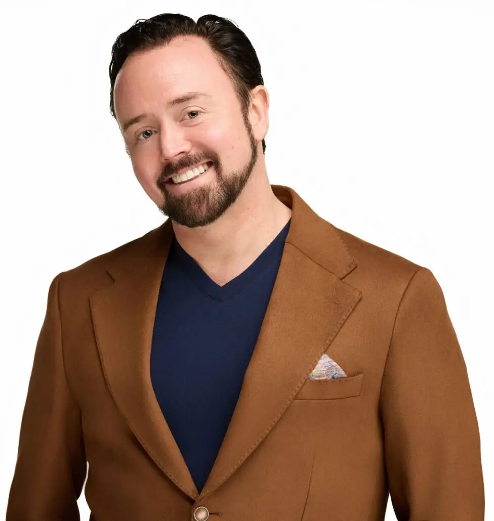 A man with short dark hair and a beard, wearing a brown blazer over a navy blue V-neck shirt, stands against a plain white background and smiles at the camera, ready to discuss the Carnival Cruise Director Schedule for Carnival Cruise 2025.