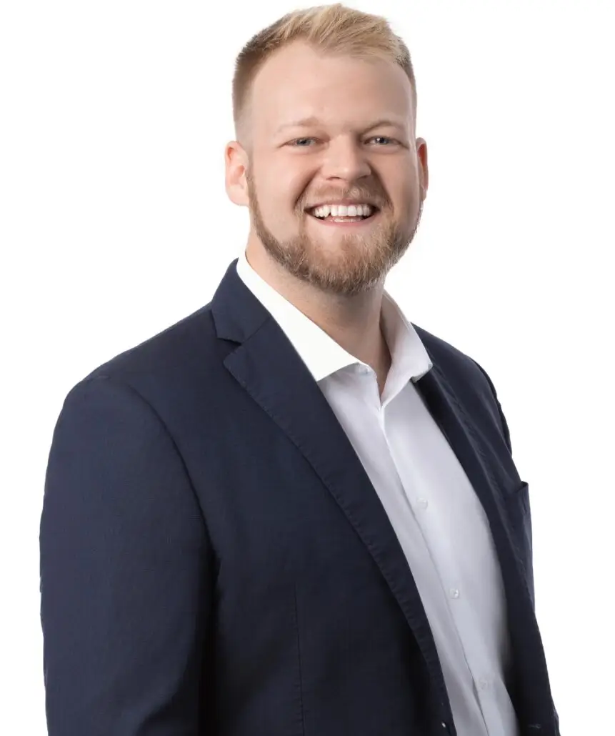 A man with short blond hair and a beard, wearing a dark blue suit jacket over a white dress shirt, smiles brightly as if ready to announce the 2025 Carnival Cruise Director Schedule, standing against a plain white background.