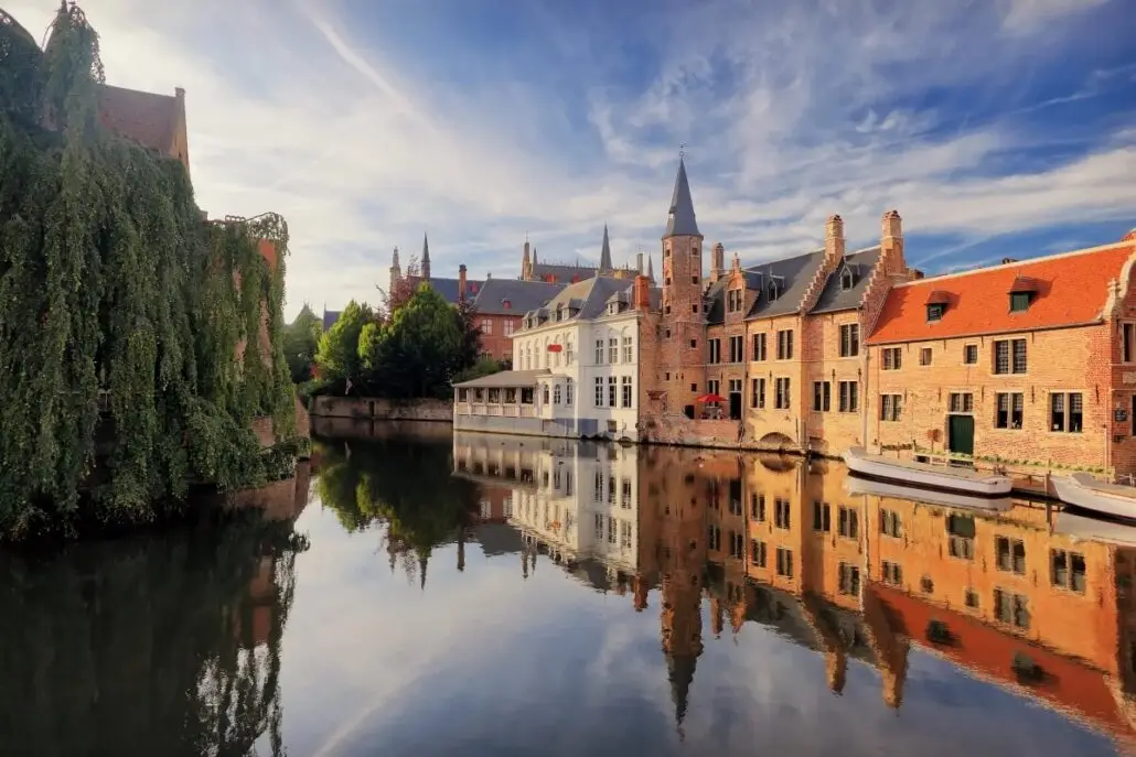 A calm river reflects historic brick buildings and a church with a spire under a partly cloudy sky; two boats, perhaps awaiting a delayed refit like those of P&O Cruises, are docked along the right side, and trees line the left bank.