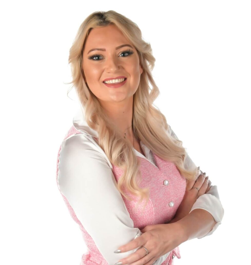 A woman with long blonde hair, dressed in a white shirt and pink sleeveless vest, stands with arms crossed and smiles at the camera—looking ready to share the 2025 Carnival Cruise Director Schedule.