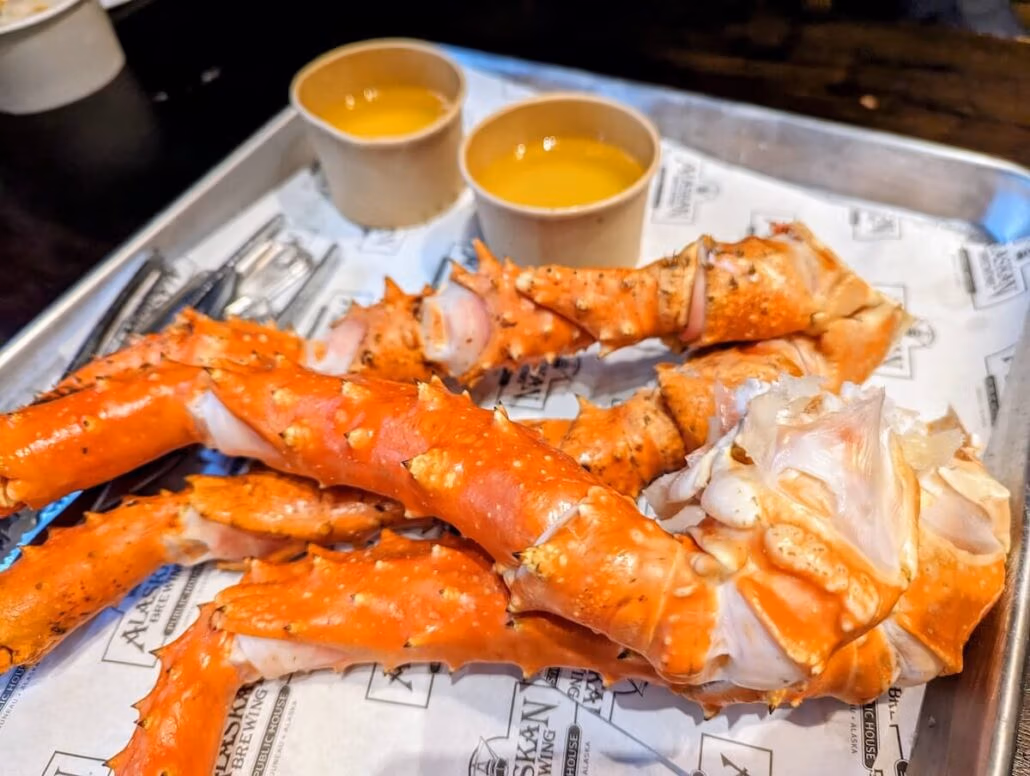 A metal tray holds several large pieces of cooked king crab legs at Alaskan Brewing Company from our cruise—with two small cups of melted butter and metal seafood utensils beside them.