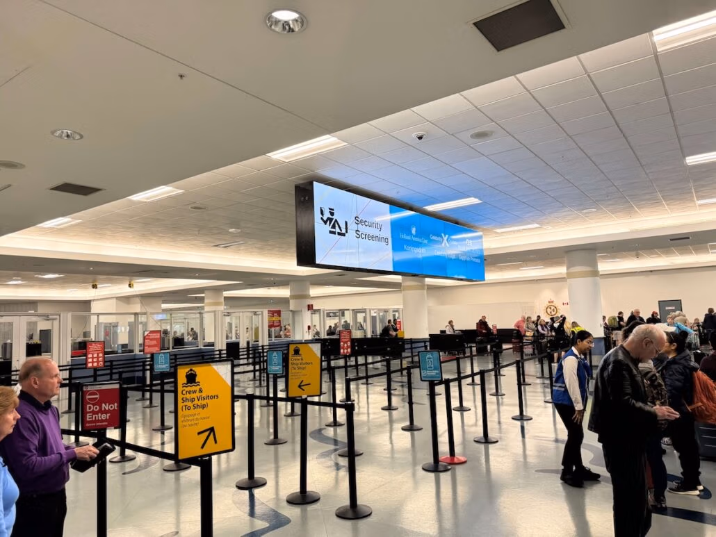People stand in line at an airport security screening area, with signs directing travelers—including those heading on a Holland America Koningsdam Alaska cruise—beneath a large overhead display reading "Security Screening." The area is brightly lit and modern.