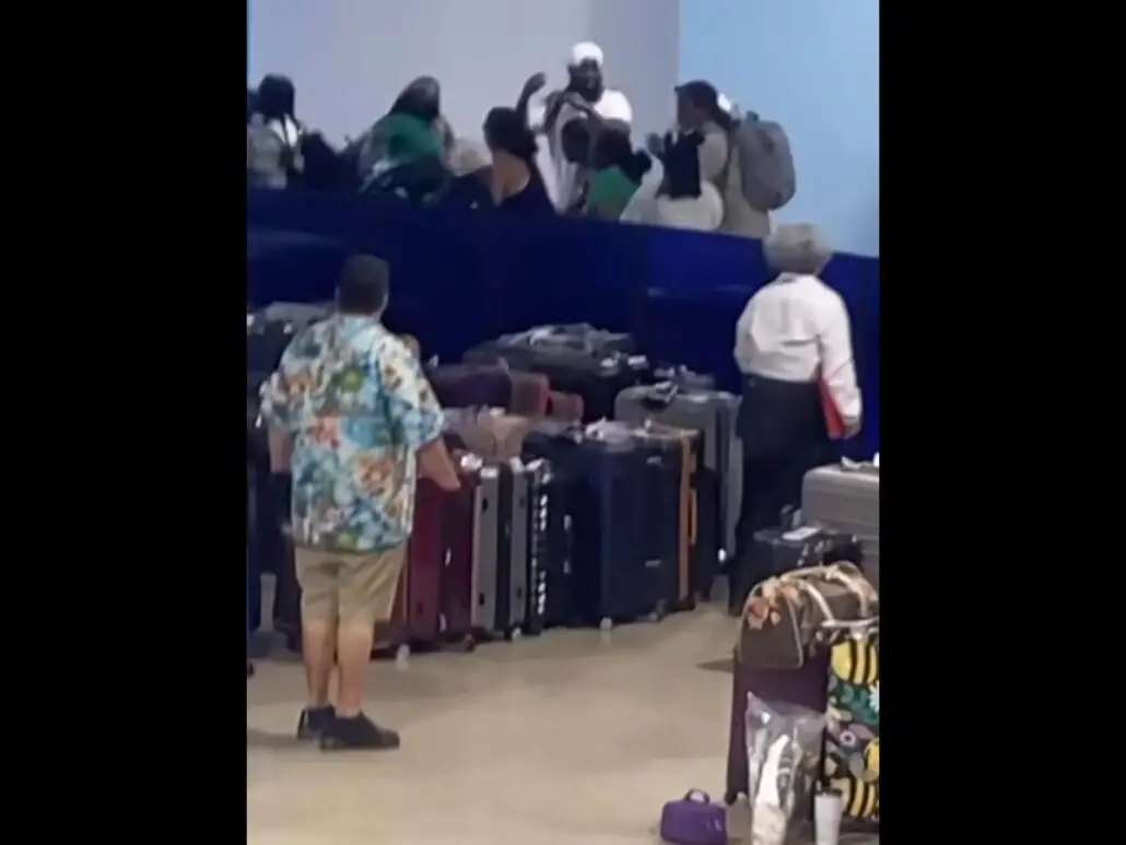 A group of people are arguing or fighting near a luggage area at a cruise terminal in Galveston. Several suitcases are lined up on the floor, while others—including Carnival Jubilee passengers—stand nearby observing the scene.
