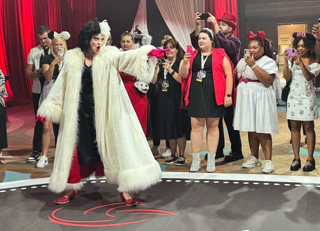 A person dressed as Cruella de Vil performs in front of a group, many snapping photos or videos. Some spectators wear costumes and dog-themed outfits. Red curtains frame the scene, evoking the magic of Disney Destiny Attractions like Hercules and The Lion King.