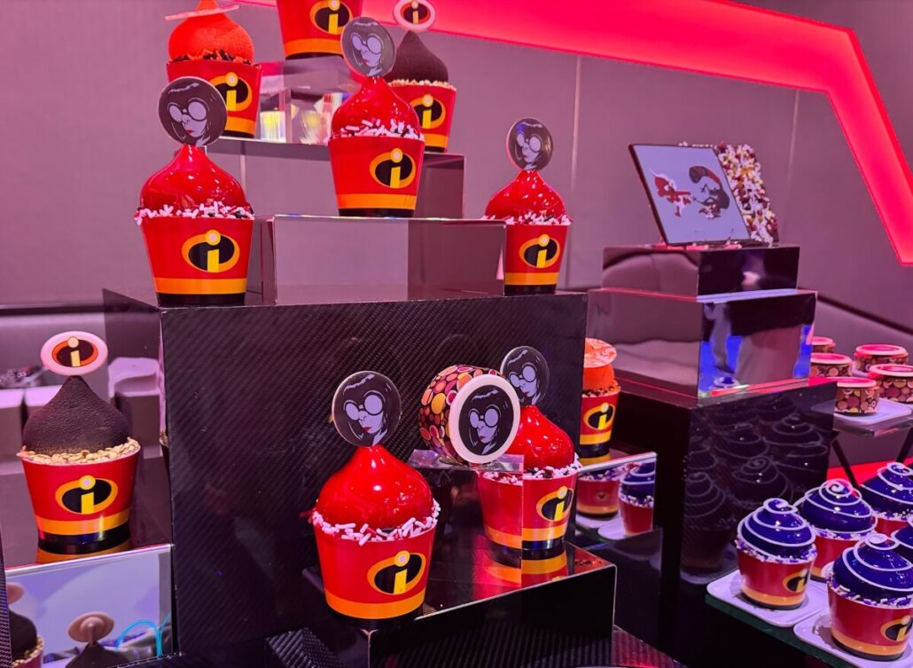 A display of cupcakes and desserts decorated with red and black “Incredibles” and “Jack-Jack” themed toppers and wrappers, set on black tiered stands under pink and purple lighting, perfect for fans of Disney Destiny Attractions.