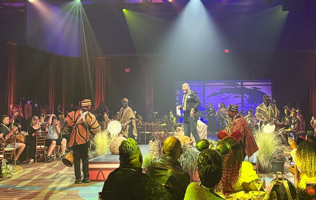 A man in a suit stands on stage speaking or performing, surrounded by drummers in traditional African attire—evoking scenes from The Lion King—with an audience seated at tables in a dimly lit venue.