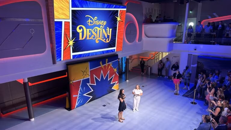 Two women stand on a stage in front of a large screen displaying “Disney Destiny” as an audience watches, surrounded by colorful decor inspired by Disney Destiny Attractions like Hercules and The Lion King in a bright indoor venue.